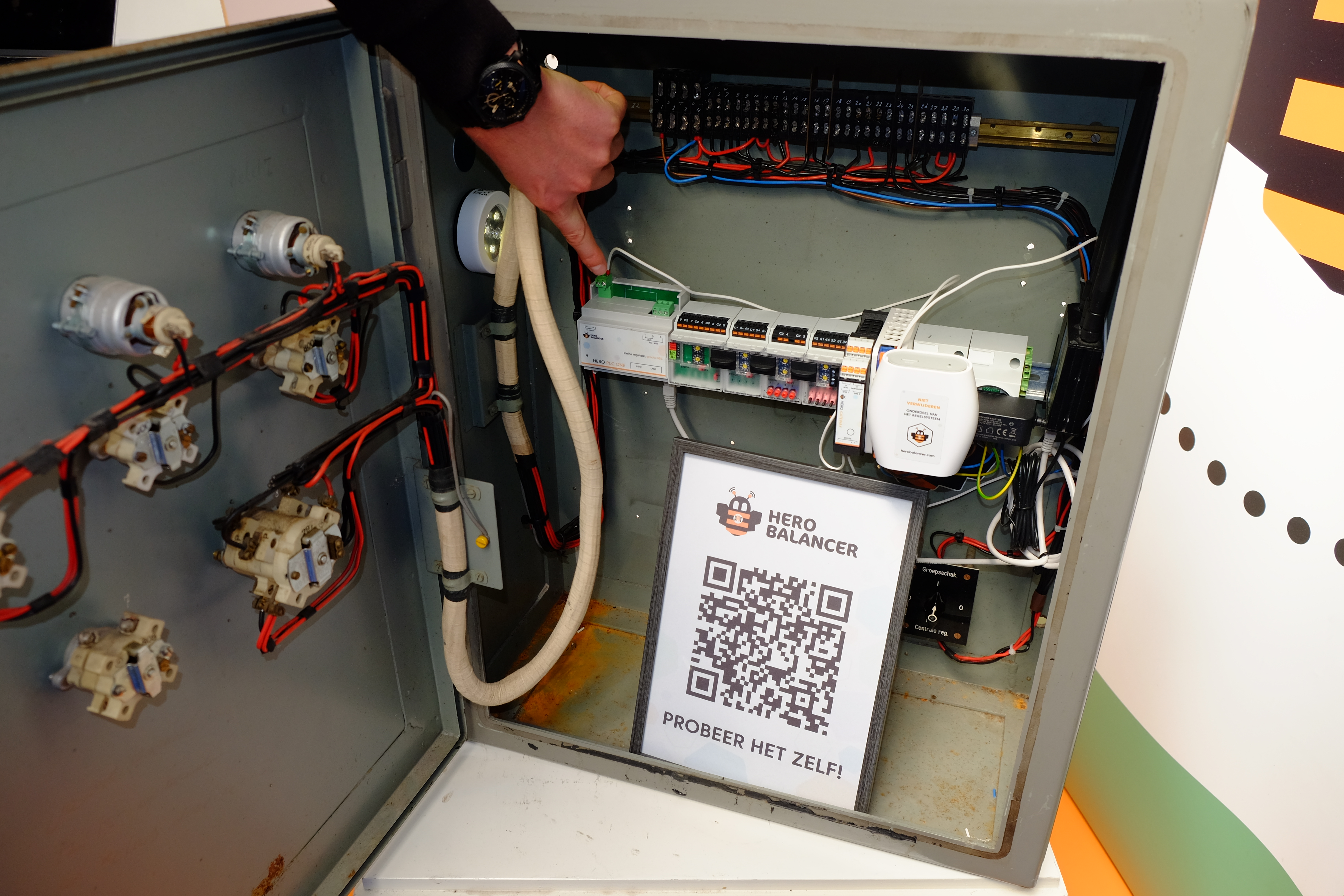 Een Hero Balancer regelkast, met links (waarnaar de vinger wijst) de regelaar en rechts de Hero Gateway en een kastje waarmee het sensorennetwerk wordt uitgelegd.